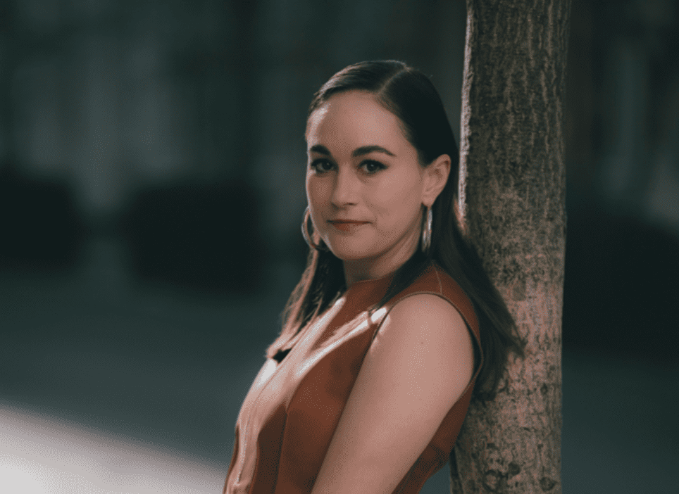 Charlotte Dales standing against tree with brown hair and orange top on