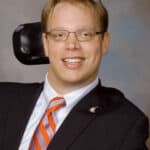 Professional headshot of Matthew wearing a black blazer and white shirt with an orange striped tie