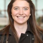 Sara Jessick, a white, young woman with long brown hair, blue eyes, and wearing a moto-style jacket, smiling at the camera.