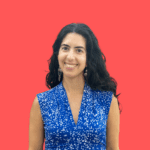 Catarina is a light-skinned Latina/Latinx woman with long dark brown wavy hair worn down. She is looking at the camera and is smiling widely. She is wearing a sleeveless blue and white top and there is a coral-colored background behind her.