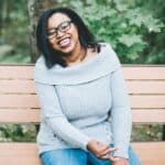 Snapshot of Keah Brown smiling, sitting on bench wearing glasses and gray top
