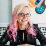 A white woman with blonde and pink hair is smiling and wearing glasses and black leather jacket.