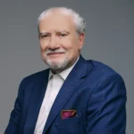 John Kemp, a white man with white hair and beard is smiling and wearing a blue checked pattern suit with burgundy handkerchief and white button down shirt.