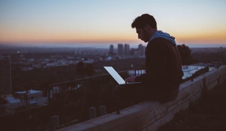A man working on a laptop outside with the sun setting.