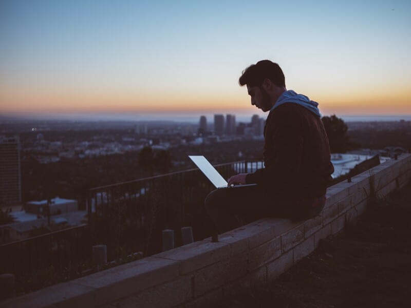 A man working on a laptop outside with the sun setting.